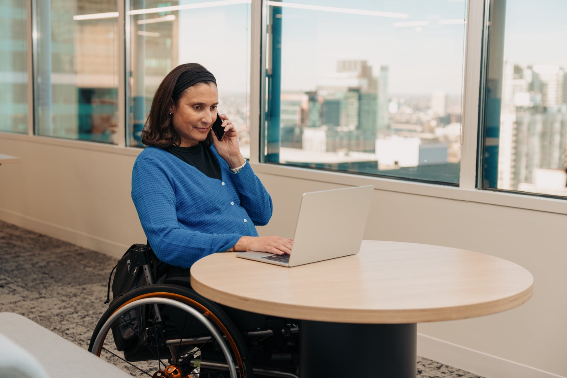 A sideview of a woman in an office environment who is on a manual wheelchair talking on the phone while resting her other hand on her laptop 