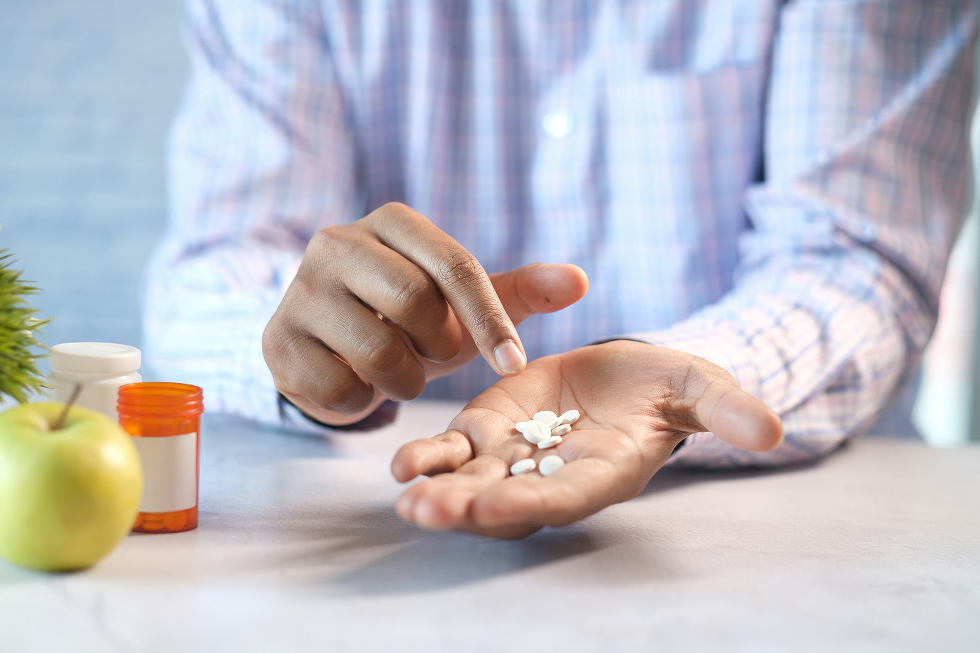 A front view of a man wearing a plaid shirt holding several white pills in one hand while pointing to them with the other hand. In front of them, there are pill bottles and an apple