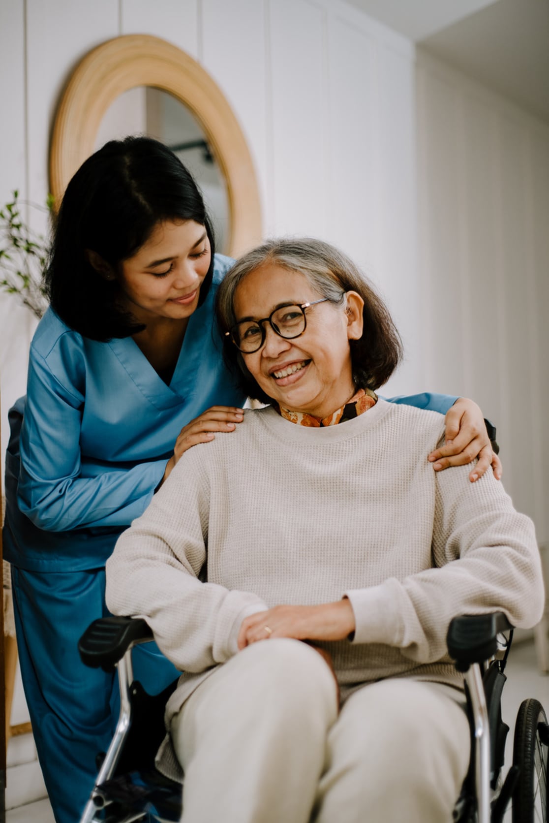 A front view of someone smiling while sitting on a manual wheelchair. Another person in scrubs is crouched down and resting their hands on the shoulders of the seated person