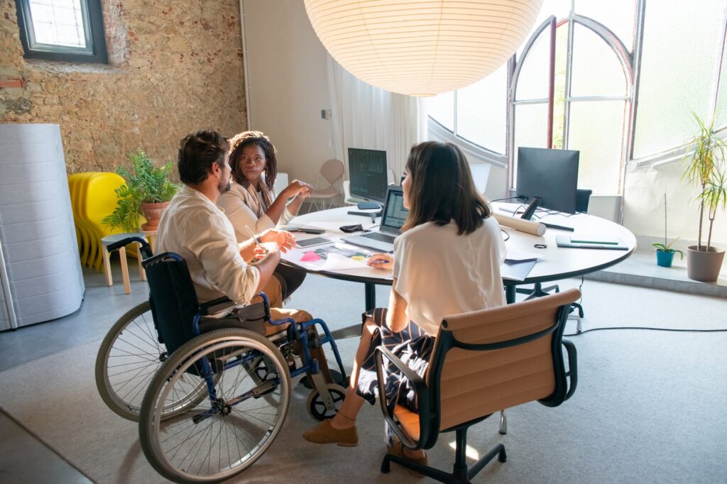 A group of three people talking while sitting around a desk that has laptops, desktop screens, and papers on it.  There is side view of someone sitting on a manual wheelchair talking to two people sitting on chairs on the opposing sides