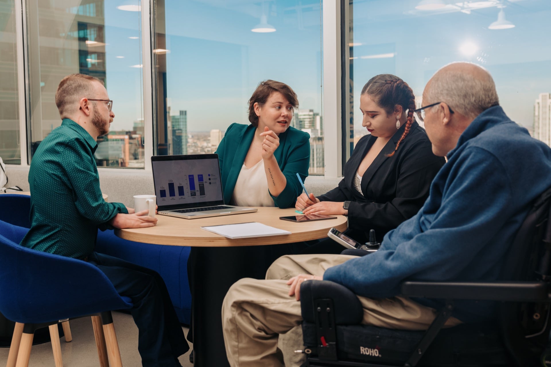 A front and side view of three people who seated around a round table in a modern office with a cityscape view through large windows. One woman is speaking while pointing to a laptop displaying charts and graphs another woman is taking notes, while the third person who is in a power wheelchair is listening attentively
