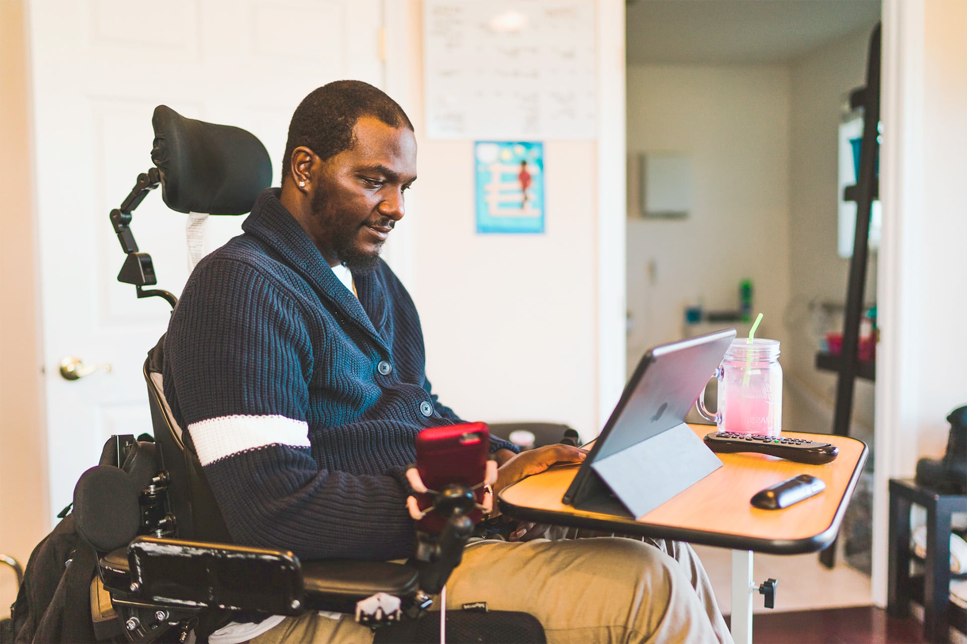 A sideview of a man in a wheelchair using a tablet and smartphone at a desk. 