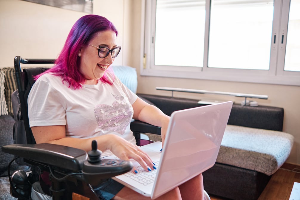 A sideview of a woman sitting in a power wheelchair using a laptop near a large window