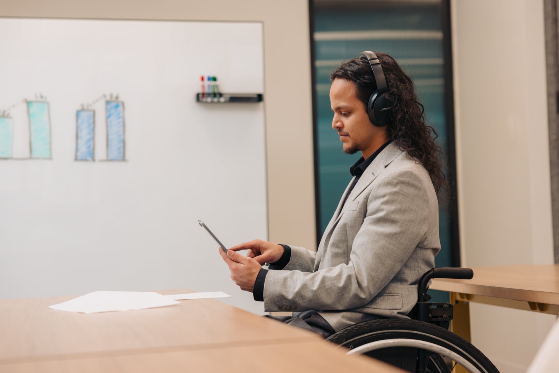A sideview of a man in a wheelchair wearing headphones, holding a tablet, seated at a desk in an office setting with a whiteboard in the background