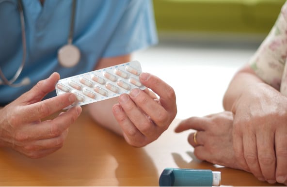 A front, close-up view of a healthcare provider wearing scrubs and a stethoscope, holding a pill blister pack. Next to them is a side close-up view of a person’s hands resting on the table next to an inhaler