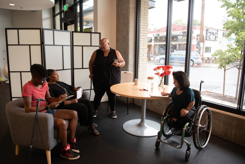 A front view of four people gathered in a room that has two chairs and a table. One person is in a manual wheelchair near the table taking notes, one is standing with a cane in their right hand, one is seated with a cane leaning on their chair and they are completing a chart, and one is seated next to them holding a to-go cup and a stack of papers