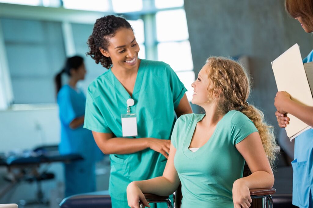 A front view of a healthcare provider wearing scrubs, standing and talking to a patient who is seated in a wheelchair. Both people are looking at each other and smiling