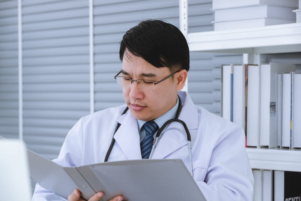 A front view of a healthcare provider wearing eyeglasses, a white lab coat, and a stethoscope around their neck reviewing a medical chart
