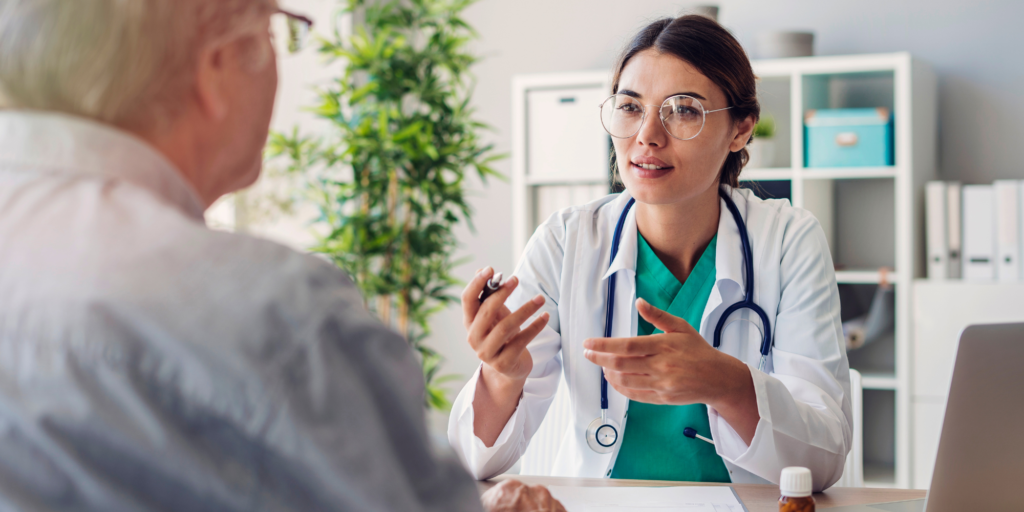 A front view of a healthcare provider wearing a white lab coat and a stethoscope around their neck speaking to a back view of a person in an office setting