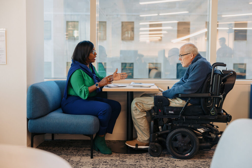 A sideview of two people sitting at a table with papers in front of them. One person is speaking while the other person who is on a power chair is listening.