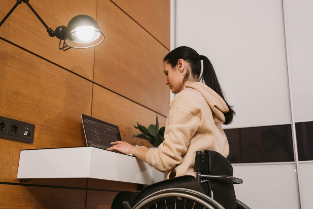 A sideview of a woman in a wheelchair sitting in front of a desk. She is typing on her laptop